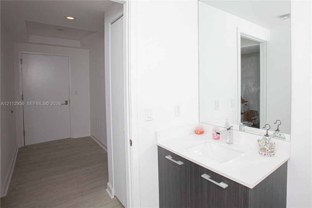 Modern vanity featuring a white composite countertop, integrated rectangular sink, polished chrome faucet, and dark wood-finish cabinetry with metallic pulls, paired with a full-height wall mirror and wood-finish flooring