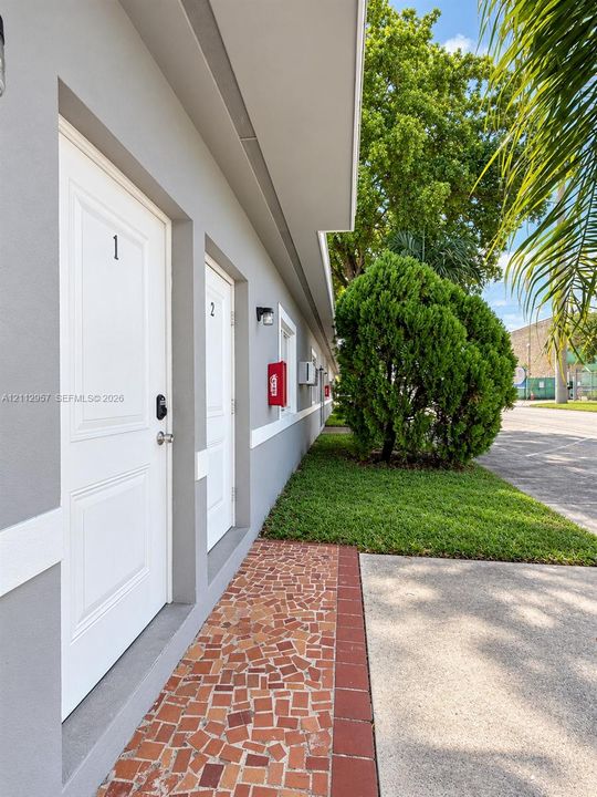 Exterior facade featuring smooth grey stucco, white-trimmed door frames, mosaic tile walkway with brick edging, and manicured landscaping