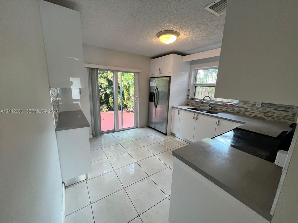 Kitchen featuring white high-gloss cabinetry, grey solid-surface countertops, horizontal tile backsplash, stainless steel refrigerator, and large-format floor tiles