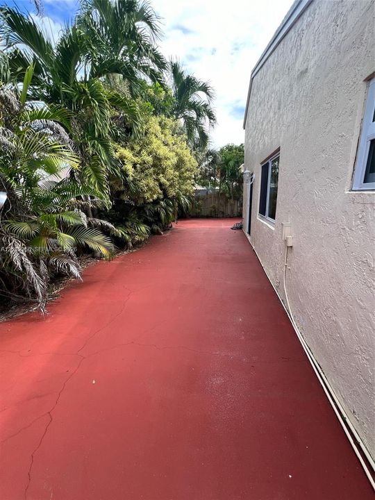 Exterior side yard featuring a red-finished concrete surface, stucco siding, and mature palm landscaping