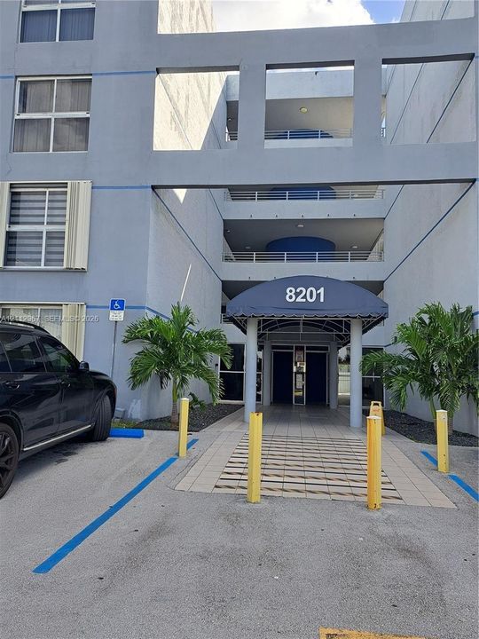Multi-story building exterior featuring a covered entryway with an arched awning, paved pedestrian path, and balconies with metal railings