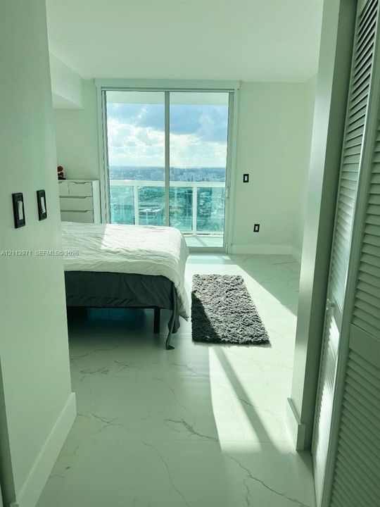 Large-format white tile flooring with veining, floor-to-ceiling glass sliding door, private balcony, and louvered closet doors