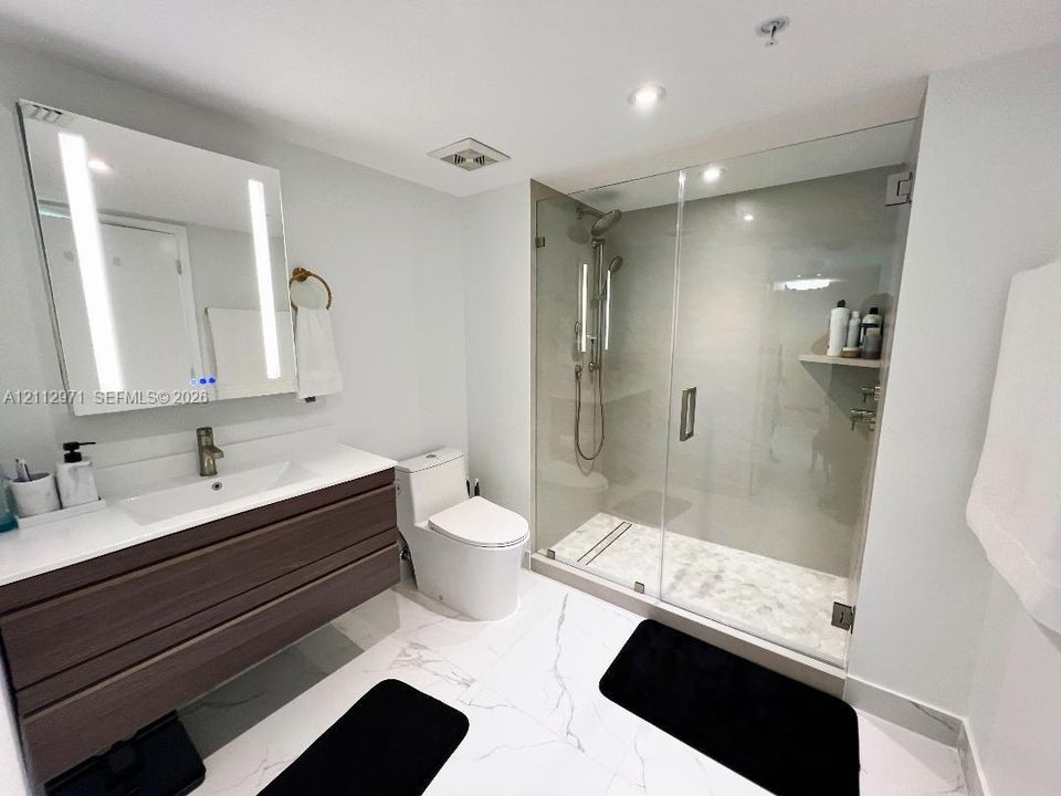 Modern vanity with a light-integrated mirror, dual-drawer wood-finish cabinetry, a glass-enclosed shower with a handheld fixture, and white stone-patterned tile flooring