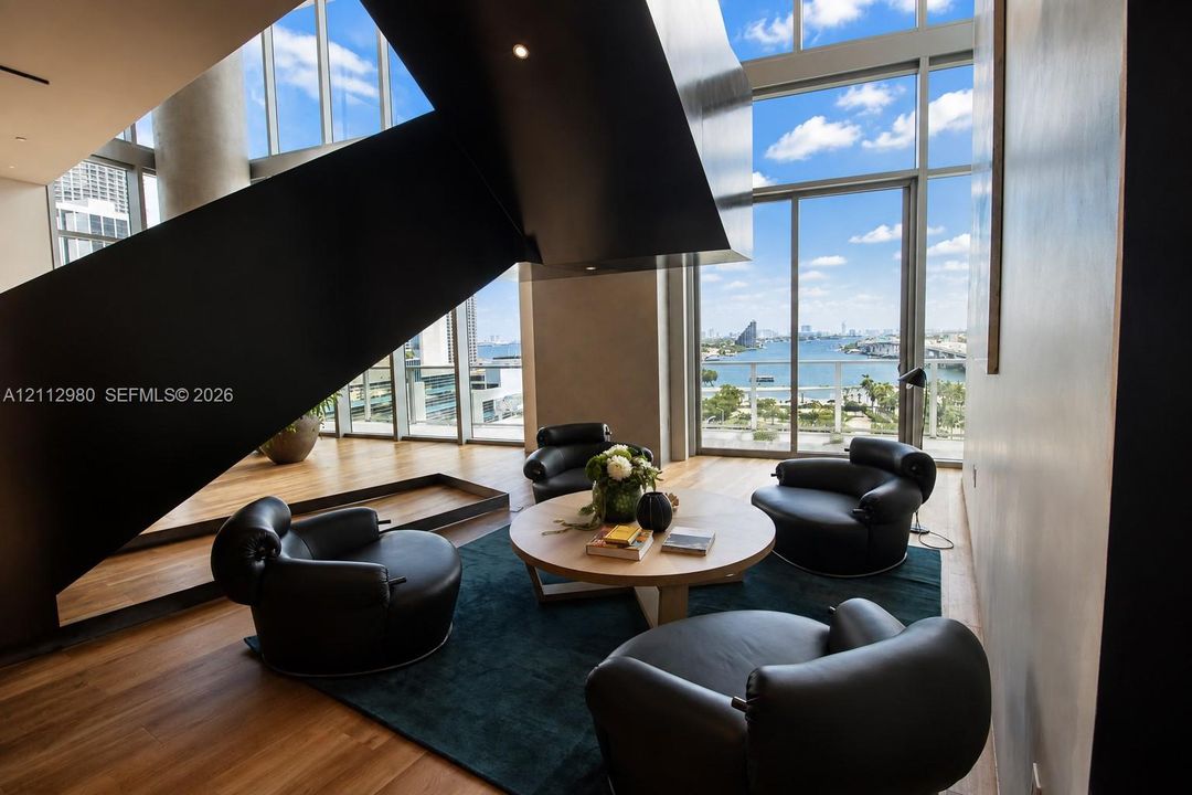Floor-to-ceiling glass walls, sculptural floating staircase, wood-finish flooring, and expansive water backdrop