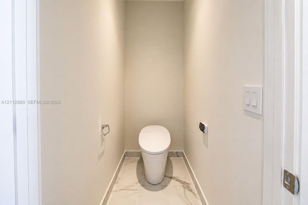 Minimalist commode alcove featuring large-format stone-patterned tile flooring, a wall-mounted flush actuator, and a sleek, one-piece toilet