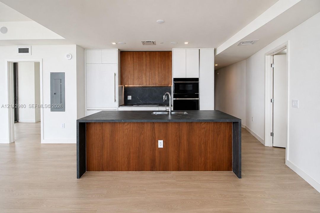 Kitchen featuring a waterfall-edge island with dark stone countertop, integrated wood-finish cabinetry, built-in wall oven, and wood-finish flooring