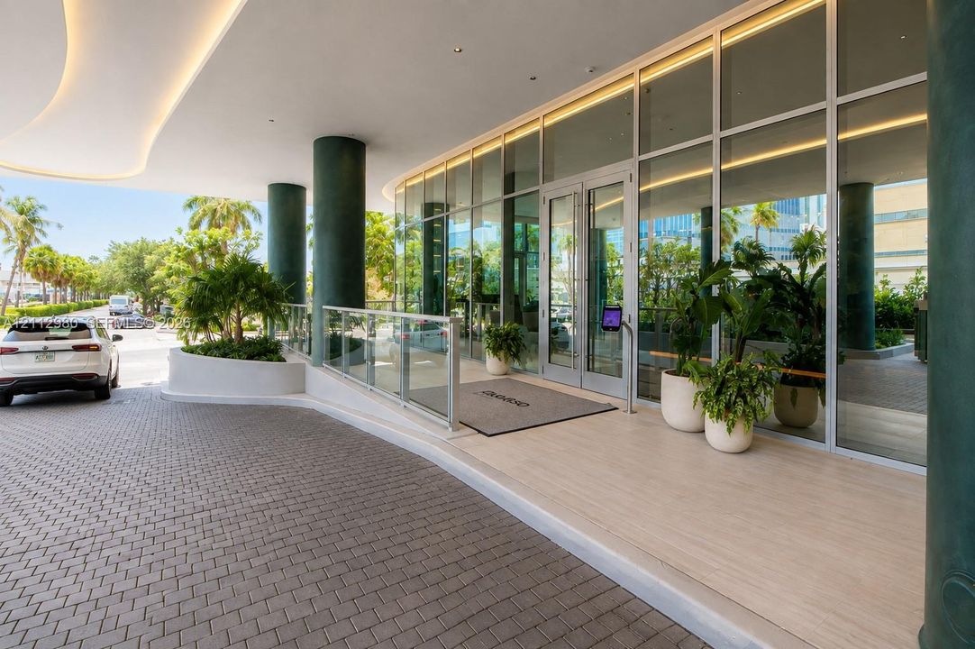 Covered motor court entrance featuring a paver driveway, floor-to-ceiling glass storefront, recessed architectural lighting, and integrated concrete planters