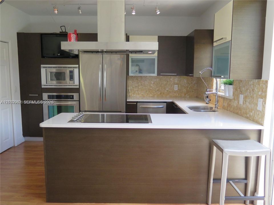 Modern kitchen featuring dark wood-finish cabinetry, white solid-surface countertops, stainless steel appliances, a flat-surface cooktop, and an island with a breakfast bar