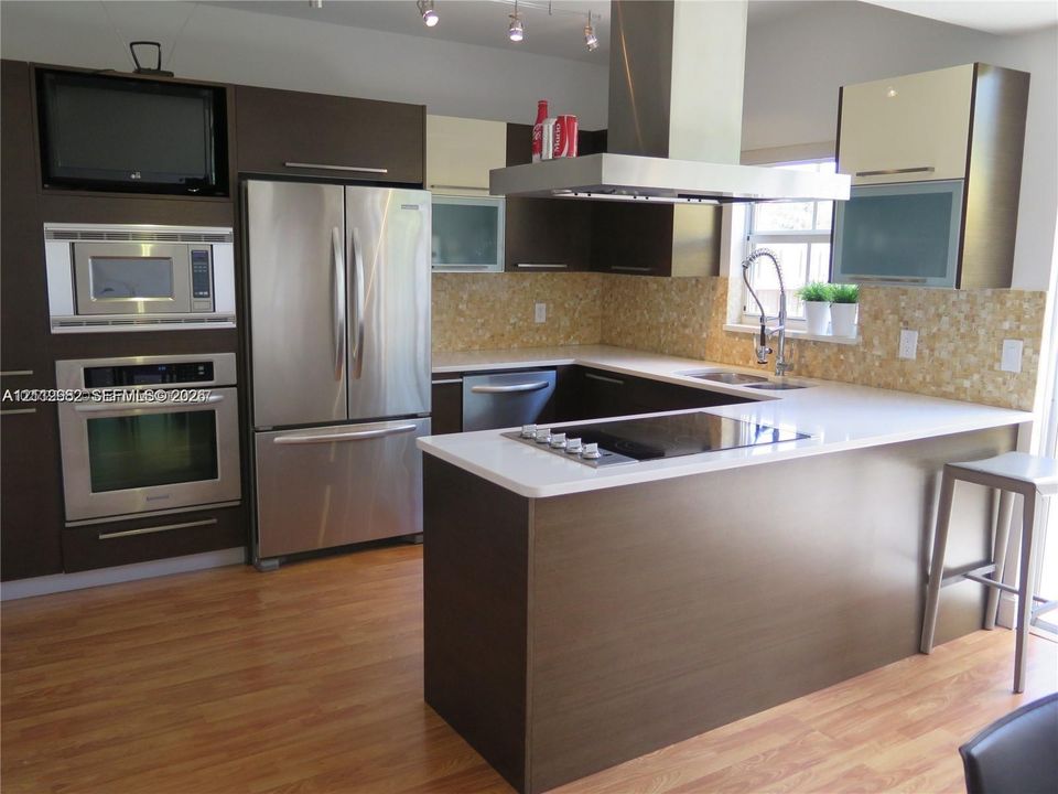 Kitchen featuring dark-finish flat-panel cabinetry, stainless steel built-in appliances, quartz countertops, a mosaic tile backsplash, and wood-finish flooring