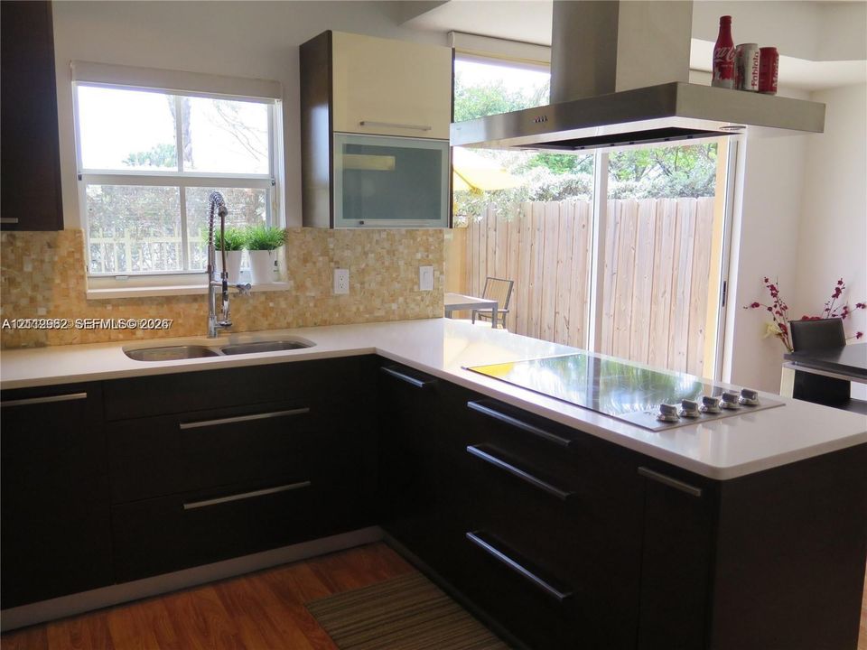 Kitchen featuring dark cabinetry with flat-panel doors, quartz countertops, a mosaic tile backsplash, a stainless steel range hood, and an inset double-basin sink with a pull-down sprayer faucet