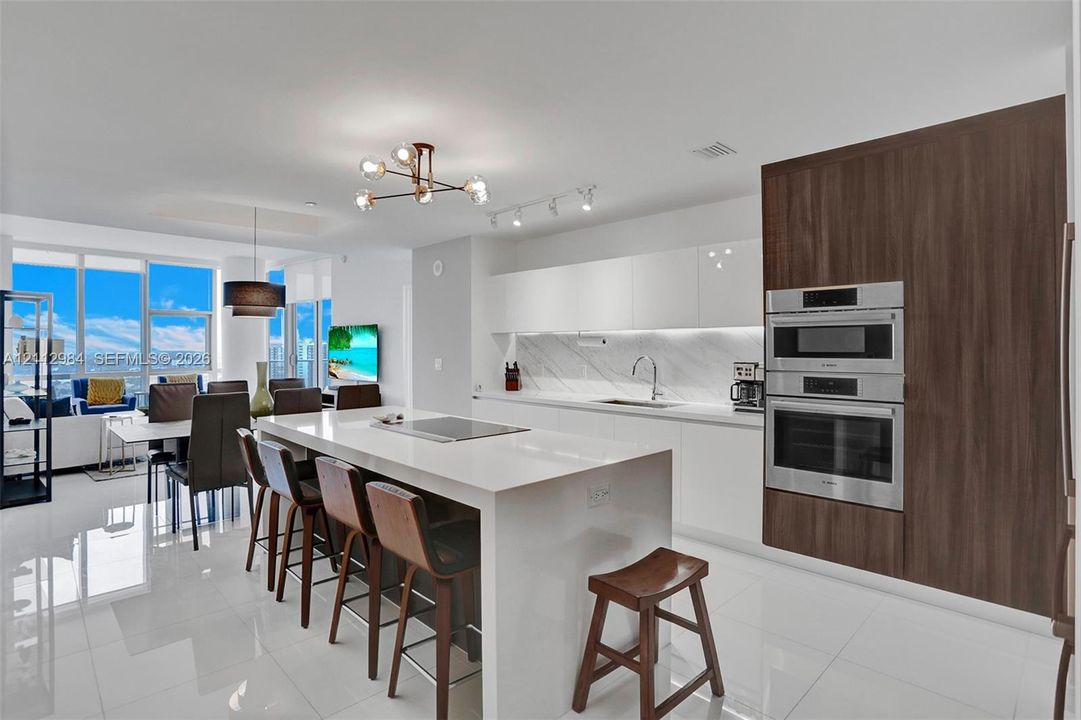 Kitchen featuring a central island with a quartz countertop, integrated cooktop, gloss-finish cabinetry, stainless steel double wall ovens, and porcelain tile flooring