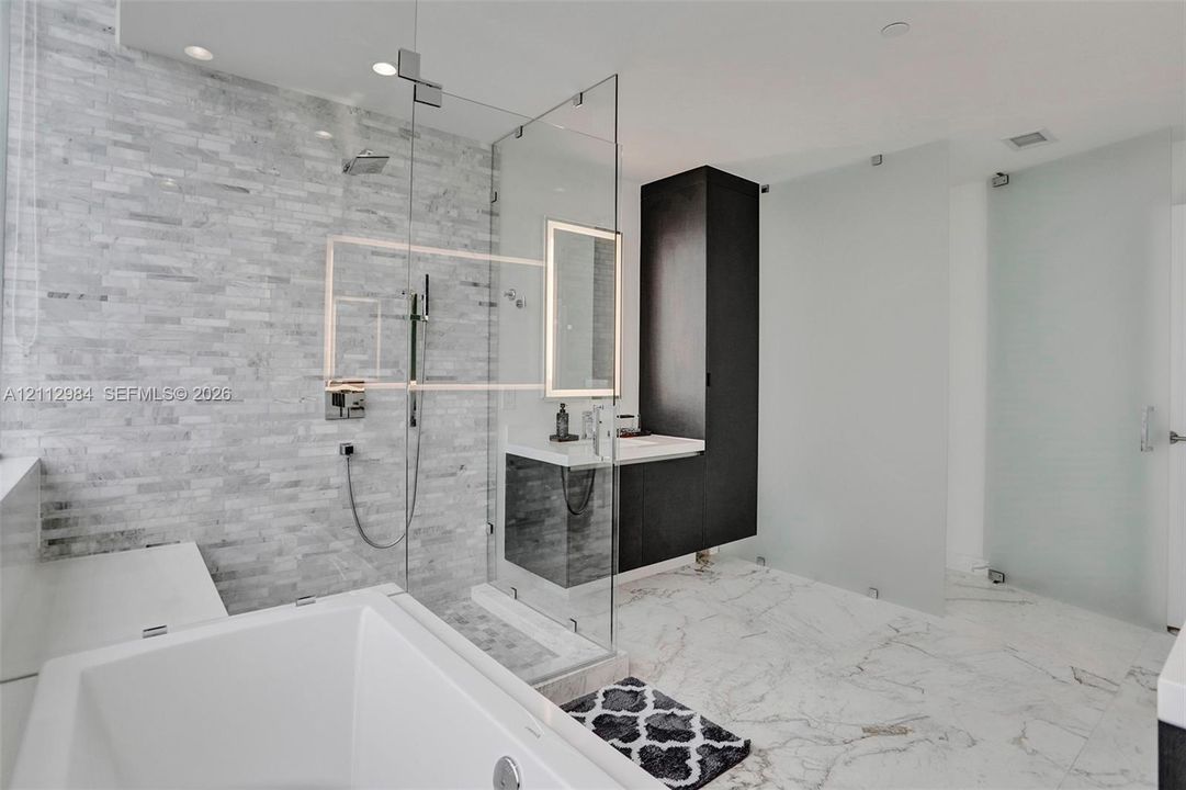 Bathroom featuring a soaking tub, stacked stone wall, frameless glass-enclosed shower, built-in floor-to-ceiling cabinetry, and light-toned stone-patterned flooring