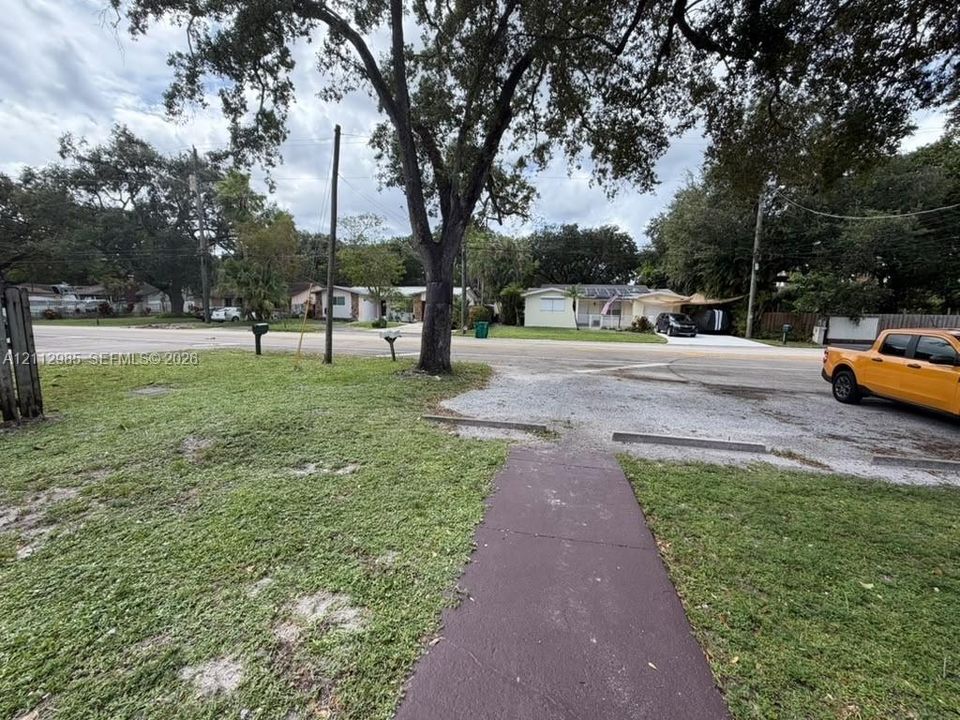 Corner lot featuring a paved walkway, gravel parking area, mature trees, and expansive lawn space