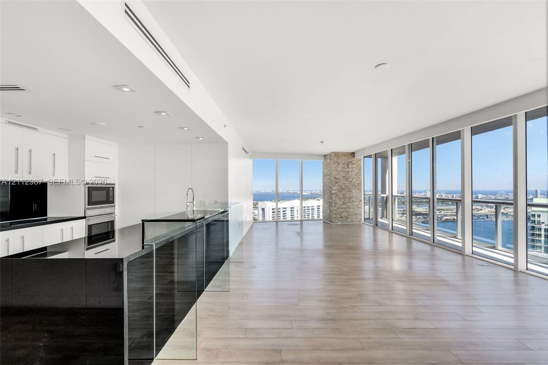 Open-concept interior featuring floor-to-ceiling windows, wood-finish flooring, a kitchen with black countertops and white cabinetry, and a stone-clad accent pillar