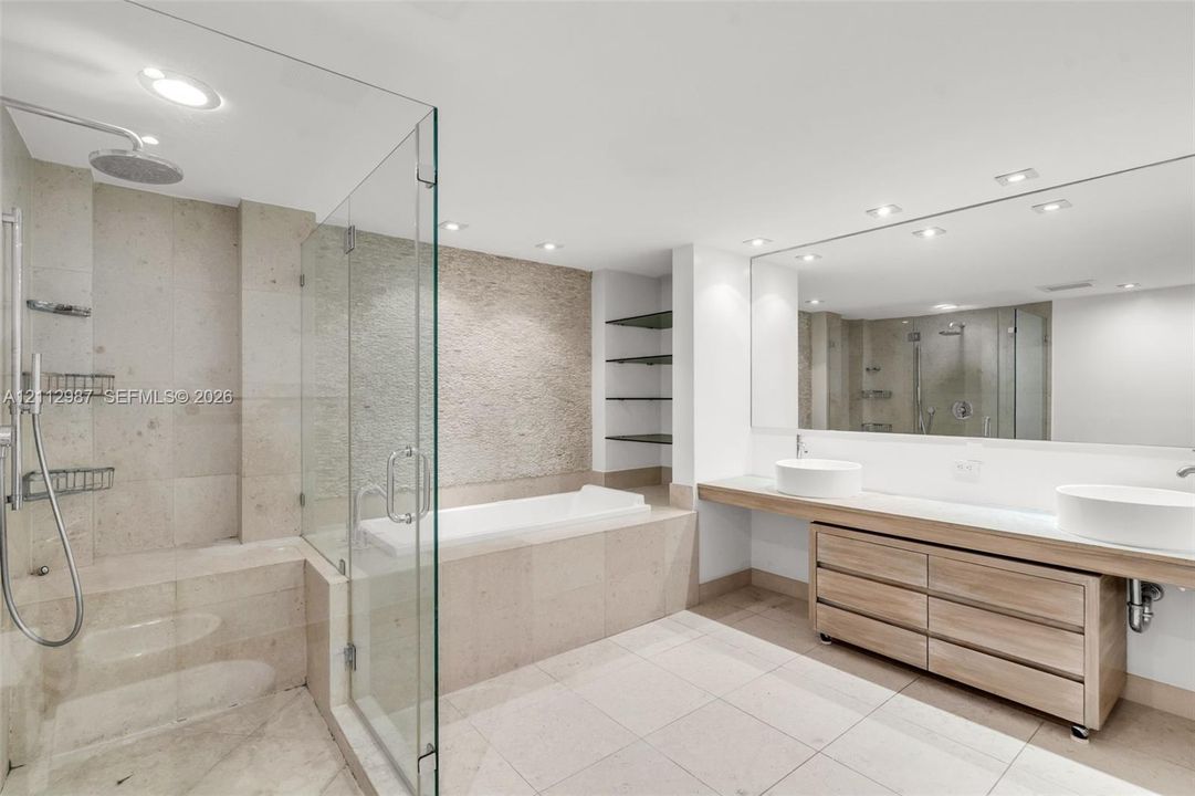 Floating wood-finish vanity with dual vessel sinks, glass-enclosed shower, built-in soaking tub, textured stone accent wall, and expansive frameless vanity mirror