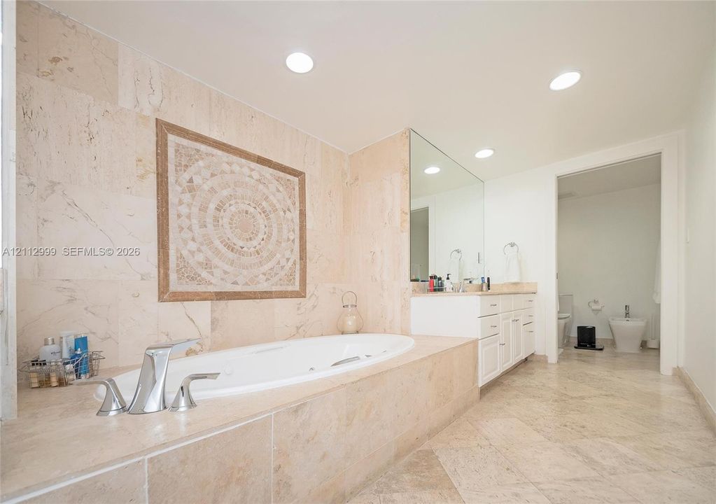 Stone-tiled soaking tub surround with decorative mosaic inlay, recessed lighting, expansive wall-mounted mirror, and stone flooring