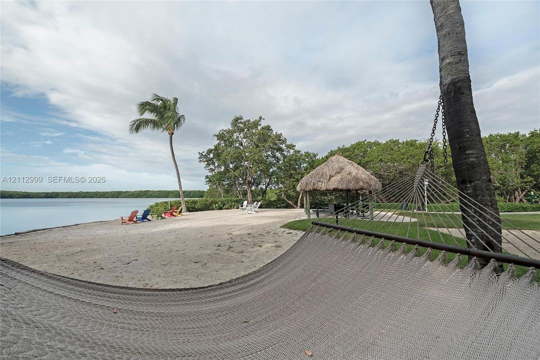 Waterfront lot featuring a sandy beach, a thatched-roof tiki hut, mature palm trees, and lush perimeter landscaping