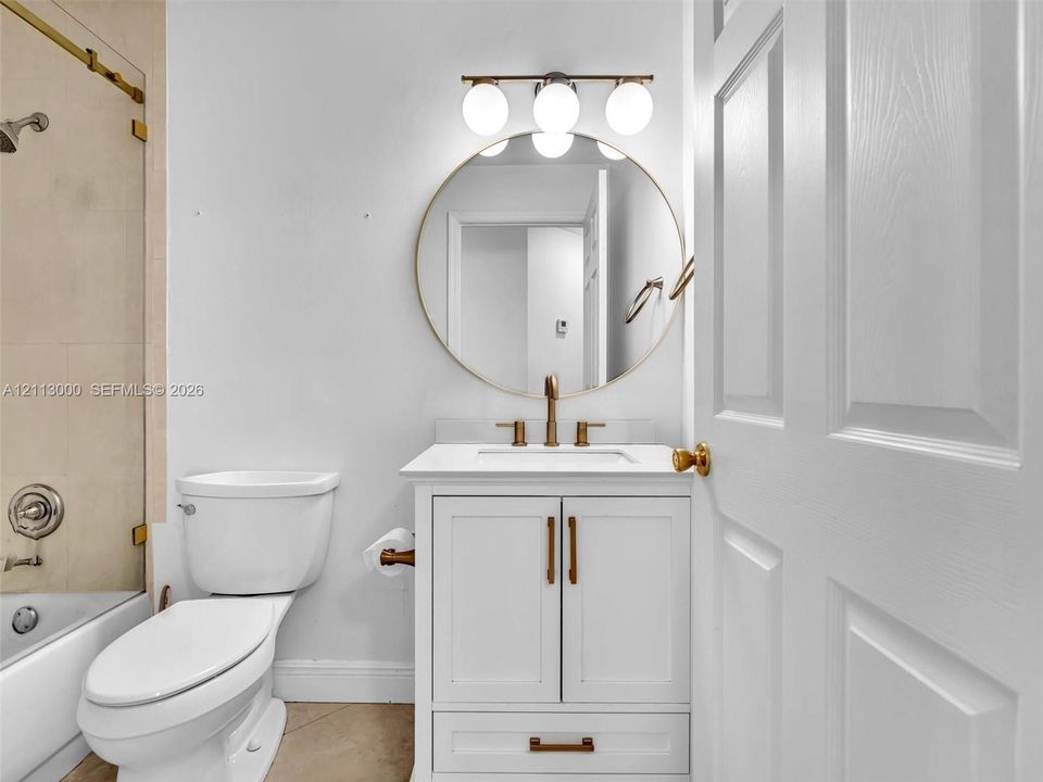 White vanity with stone countertop, brass-toned hardware, circular mirror, three-light globe vanity fixture, and glass-enclosed shower