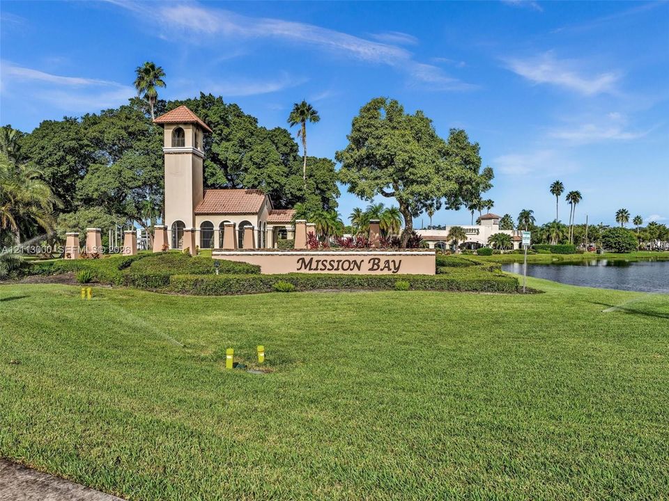 Stucco entry tower with tile roof, manicured landscaping, mature trees, and waterfront grounds