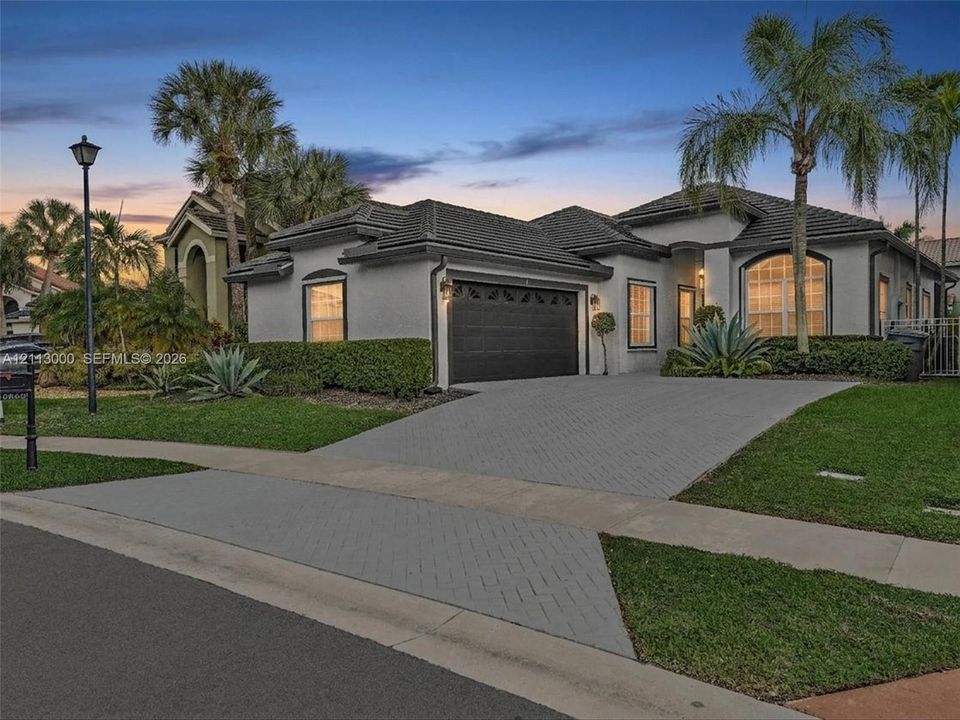 Single-story stucco exterior featuring a multi-hipped tile roof, paved driveway, arched window detailing, and mature palm landscaping