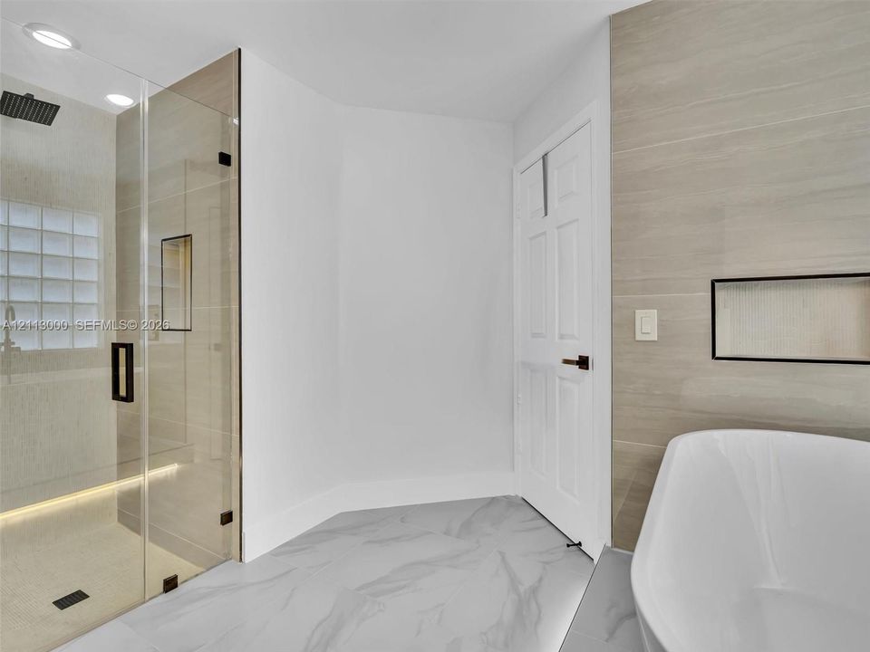 Walk-in shower with frameless glass enclosure, rainfall showerhead, and integrated recessed shelving, featuring stone-finish flooring and a freestanding soaking tub