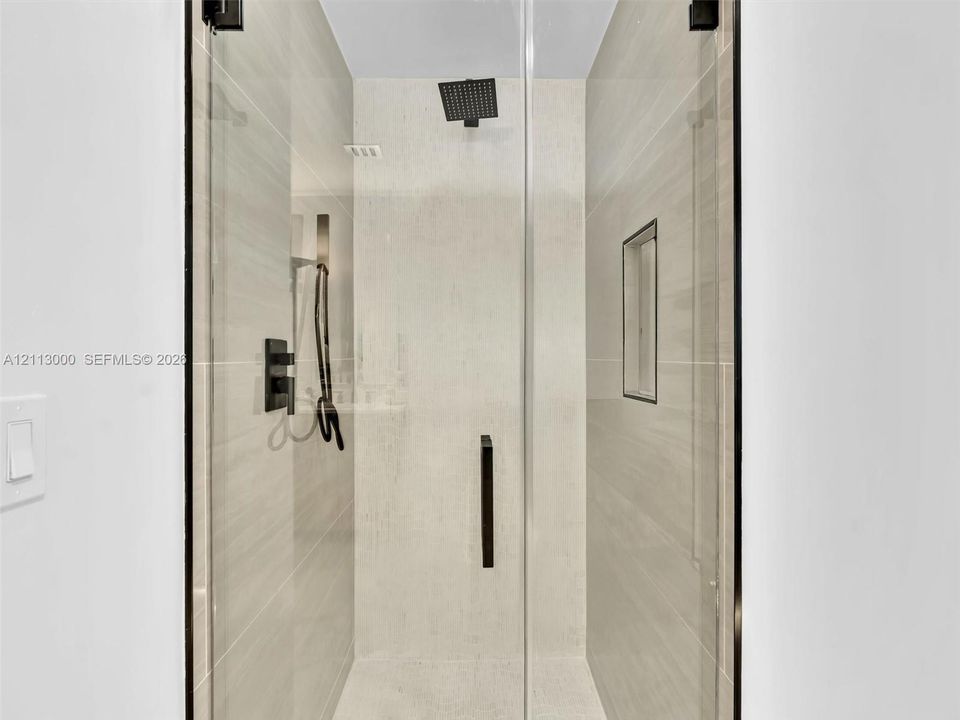 Walk-in shower featuring floor-to-ceiling tile, frameless glass enclosure, matte black rainfall showerhead with handheld sprayer, and an integrated wall niche
