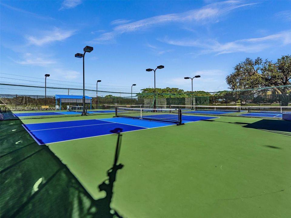 Multi-court tennis facility featuring blue and green hardcourt surfaces, perimeter chain-link fencing, and overhead court lighting