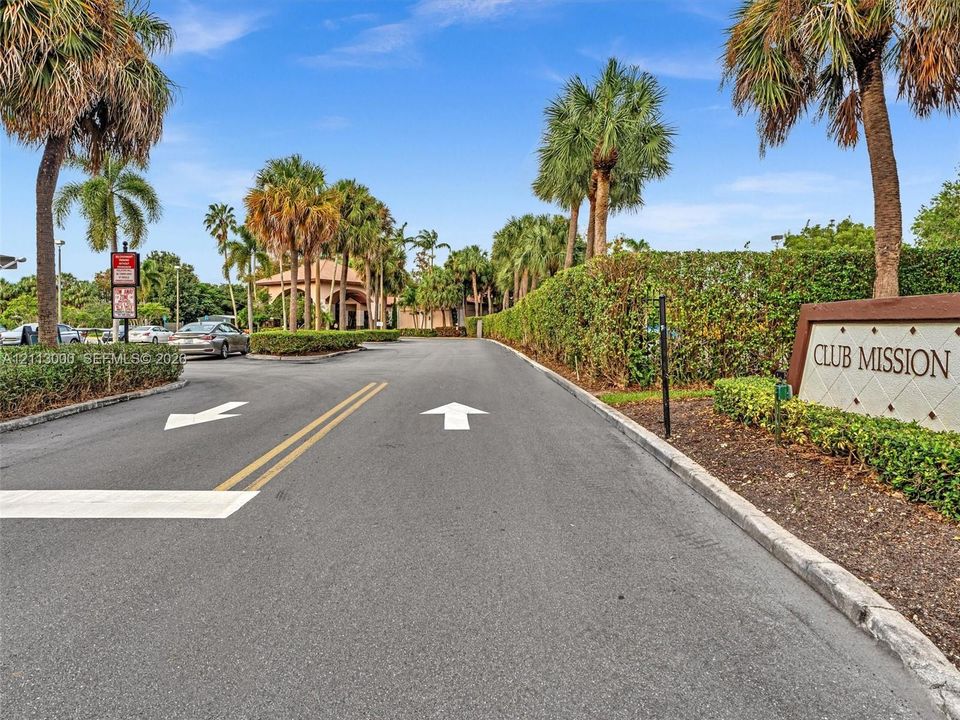 Asphalt roadway featuring paved lane markings, mature palm trees, integrated shrubbery borders, and a Club Mission monument sign