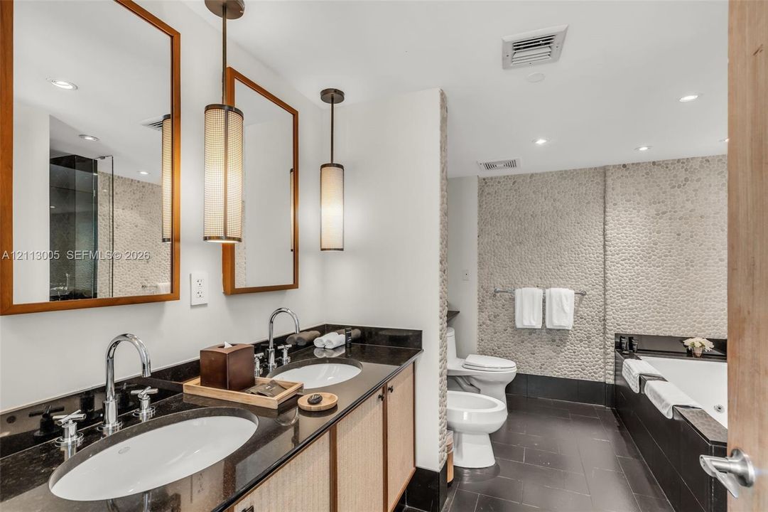 Dual-sink vanity with dark stone countertop, textured stone wall accents, recessed lighting, and modern cylindrical pendant fixtures