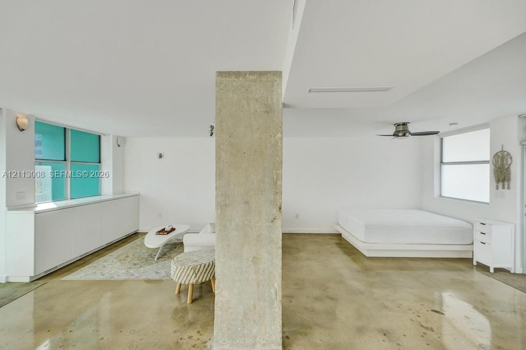 Open-concept interior featuring polished concrete flooring, a central exposed concrete structural column, high-gloss white built-in cabinetry, and a ceiling-mounted fan
