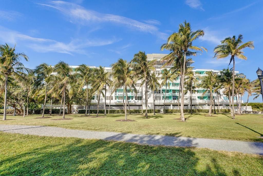 Multi-story residential building featuring white stucco facade, private recessed balconies, concrete pedestrian pathways, and mature palm landscaping situated on a expansive lawn