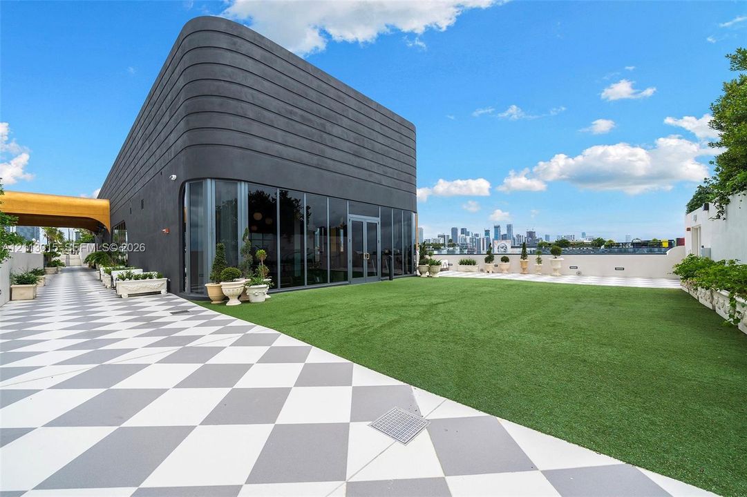 Rooftop terrace featuring large-format checkered paving, artificial turf surface, full-height glass window wall, and curved exterior structure