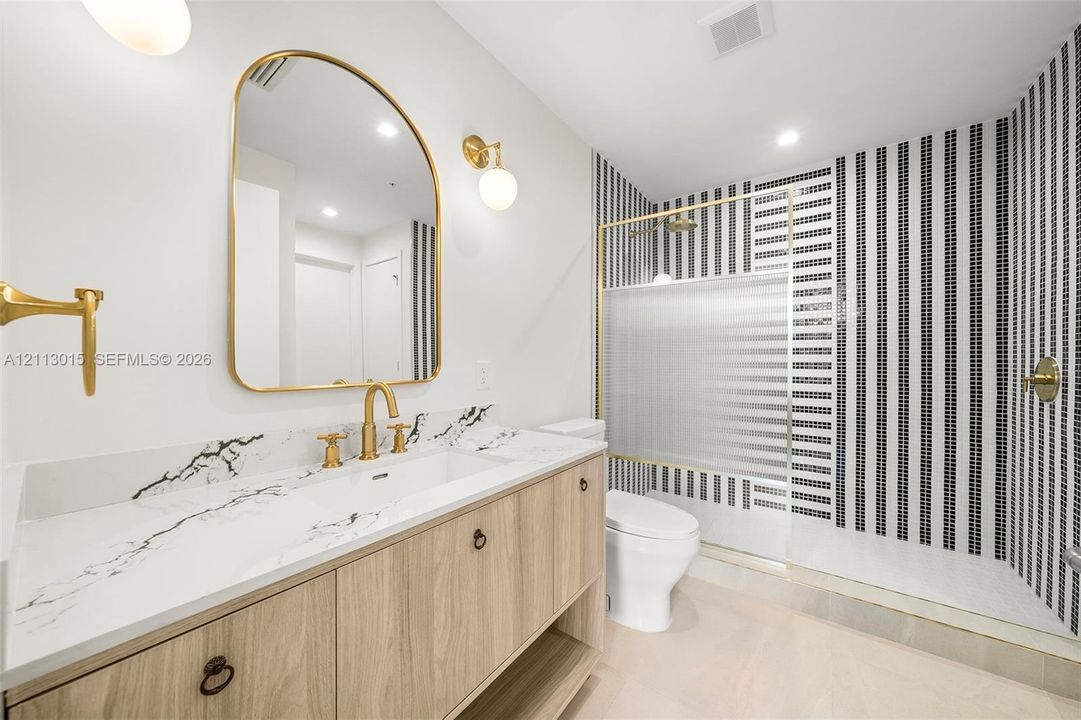 Vanity with wood-finish cabinetry, stone countertop with veining, arched wall mirror, gold-toned fixtures, and a shower enclosure featuring vertical striped mosaic tiling