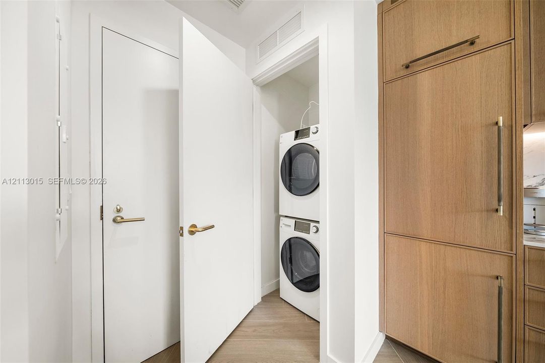 Laundry closet featuring a stacked washer and dryer, wood-finish flooring, floor-to-ceiling wood cabinetry, and satin brass hardware