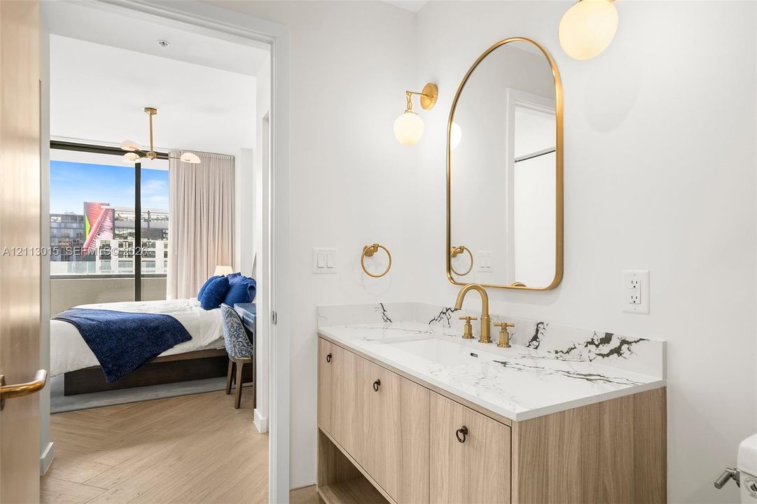 Modern vanity with a veined stone countertop and integrated sink, complemented by a brass-finished arched mirror, wall-mounted globe lighting, and light wood-finish flooring