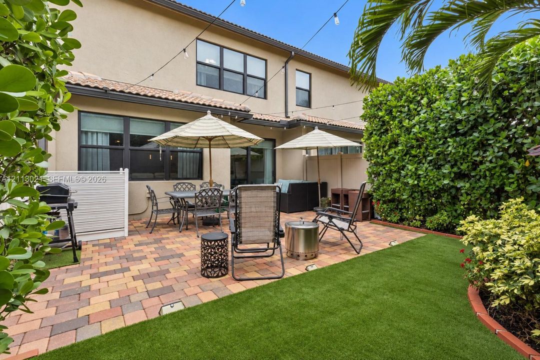 Rear patio featuring multicolored paver hardscaping, synthetic turf lawn, privacy hedging, and overhead string lighting