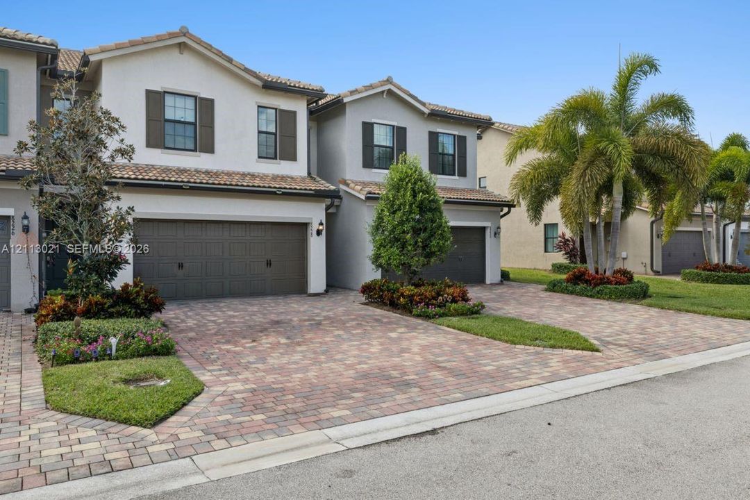 Two-story multi-family exterior featuring a tiled roof, paver driveway, attached multi-car garage, and established landscaping with palm trees