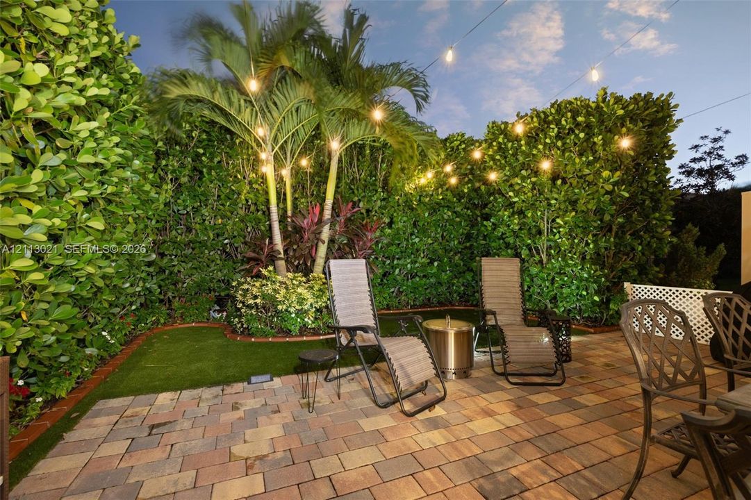 Private outdoor space featuring a paved brick patio, manicured synthetic lawn, perimeter privacy hedges, and palm trees