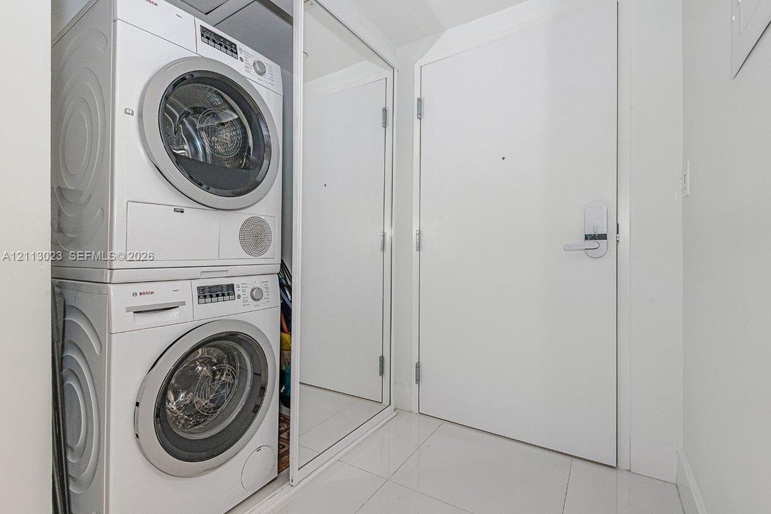 Stacked Bosch laundry center enclosed by sliding mirrored doors, set against large-format tile flooring and a solid-core door featuring electronic keypad entry hardware
