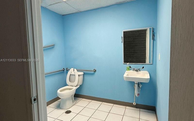 Wall-mounted porcelain sink with dual-handle faucet, white tile flooring, and metal safety grab bars