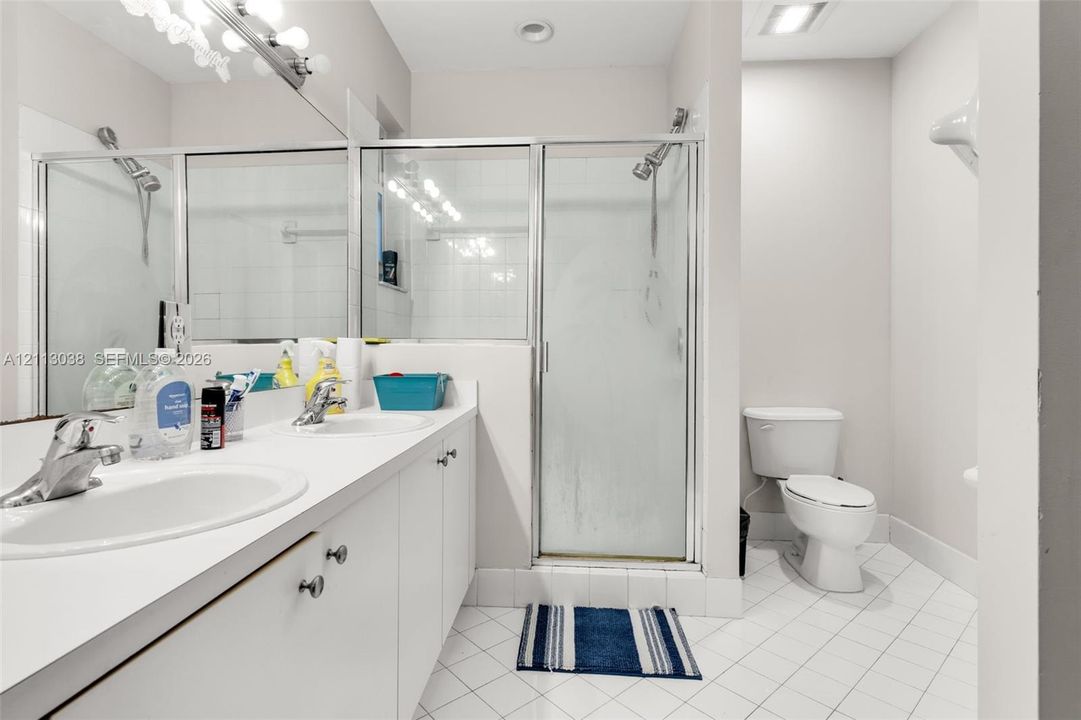 Dual-sink vanity with white cabinetry and solid-surface countertop, walk-in shower with glass enclosure, and white tile flooring