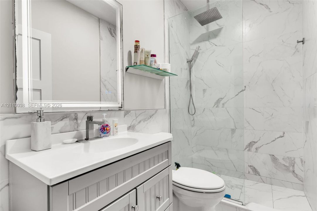 Bathroom featuring a vanity with a white countertop, an integrated rectangular sink, a LED-backlit mirror, floor-to-ceiling tiled shower surround, and a ceiling-mounted rainfall showerhead