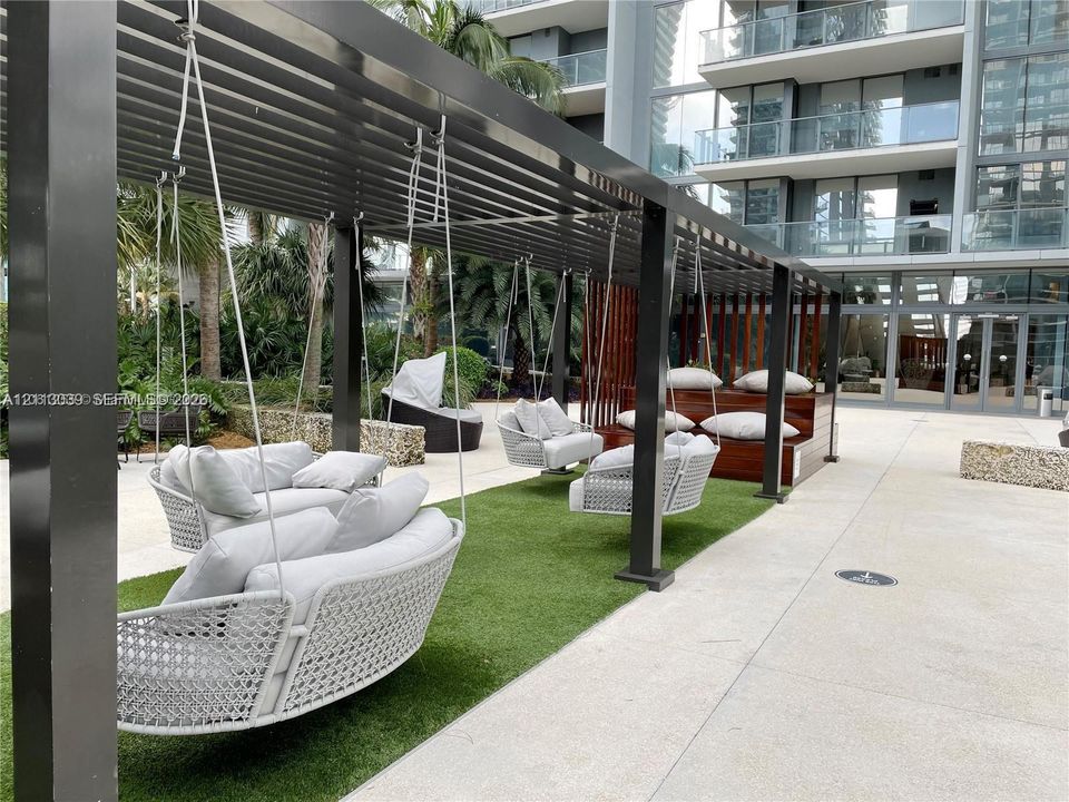 Outdoor common area featuring a black metal pergola, integrated artificial turf flooring, built-in tiered wood-tone seating, and suspended seating structures