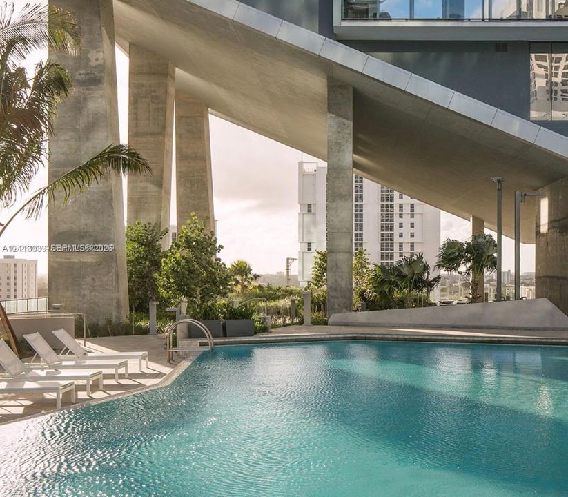 Swimming pool featuring concrete structural columns, geometric architectural overhang, paved sun deck, and tropical landscape elements