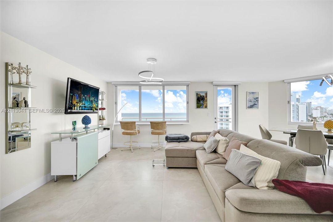 Open-concept living area featuring large-format tile flooring, a modern sculptural chandelier, floor-to-ceiling windows with coastal scenery, and a glass-paneled entry door