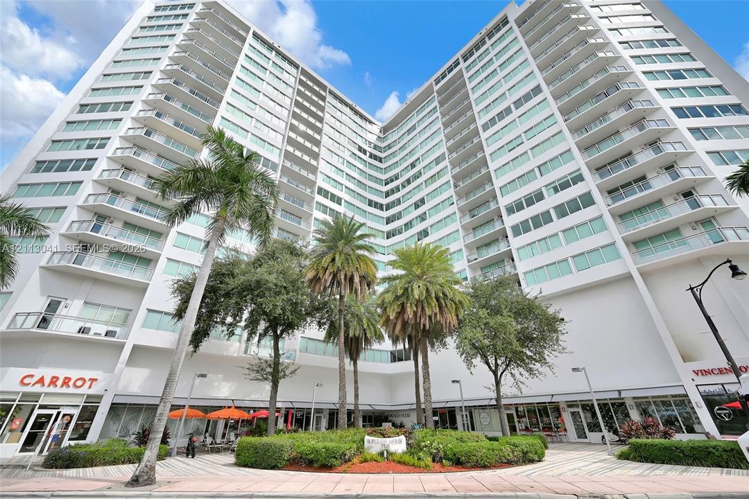 High-rise concrete exterior featuring expansive glass windows and private balconies, complemented by ground-level commercial frontage and manicured landscaping with mature palm trees