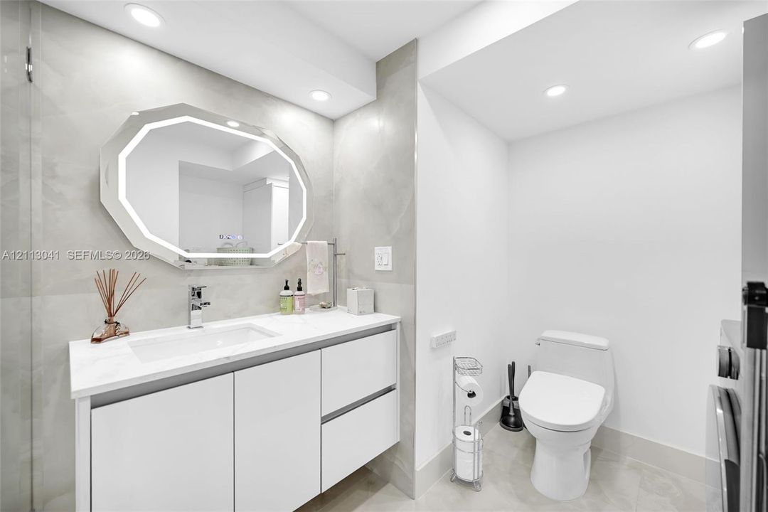 Modern floating vanity featuring white cabinetry, a rectangular sink, and a light-toned countertop, paired with an octagonal LED-backlit mirror and large-format wall tile