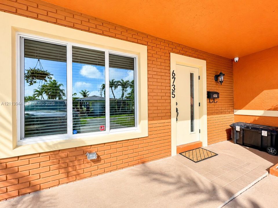 Exterior entrance featuring painted brick masonry, large picture window with wide-frame casing, solid-core door with glass inset, and integrated wall-mounted light fixture