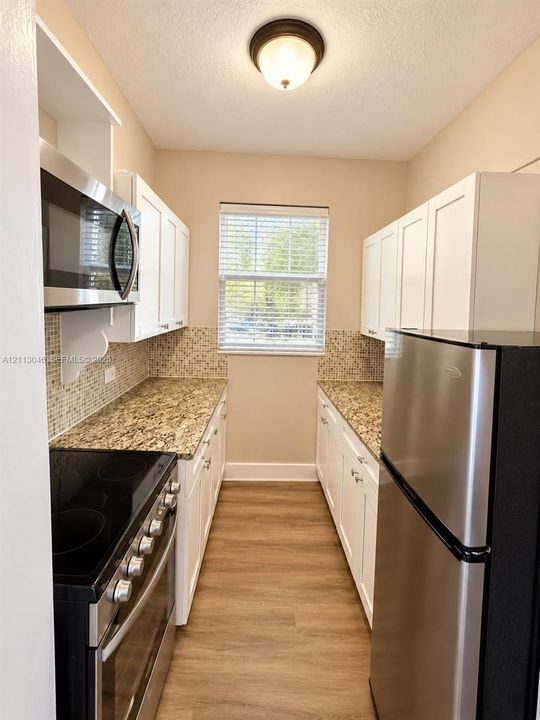 Galley kitchen featuring white shaker-style cabinetry, stone countertops, mosaic tile backsplash, stainless steel appliances, and wood-finish flooring