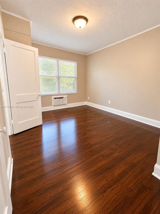Room interior featuring dark wood-finish flooring, neutral wall paint, a flush-mount ceiling light, a white paneled door, and a wall-mounted climate control unit